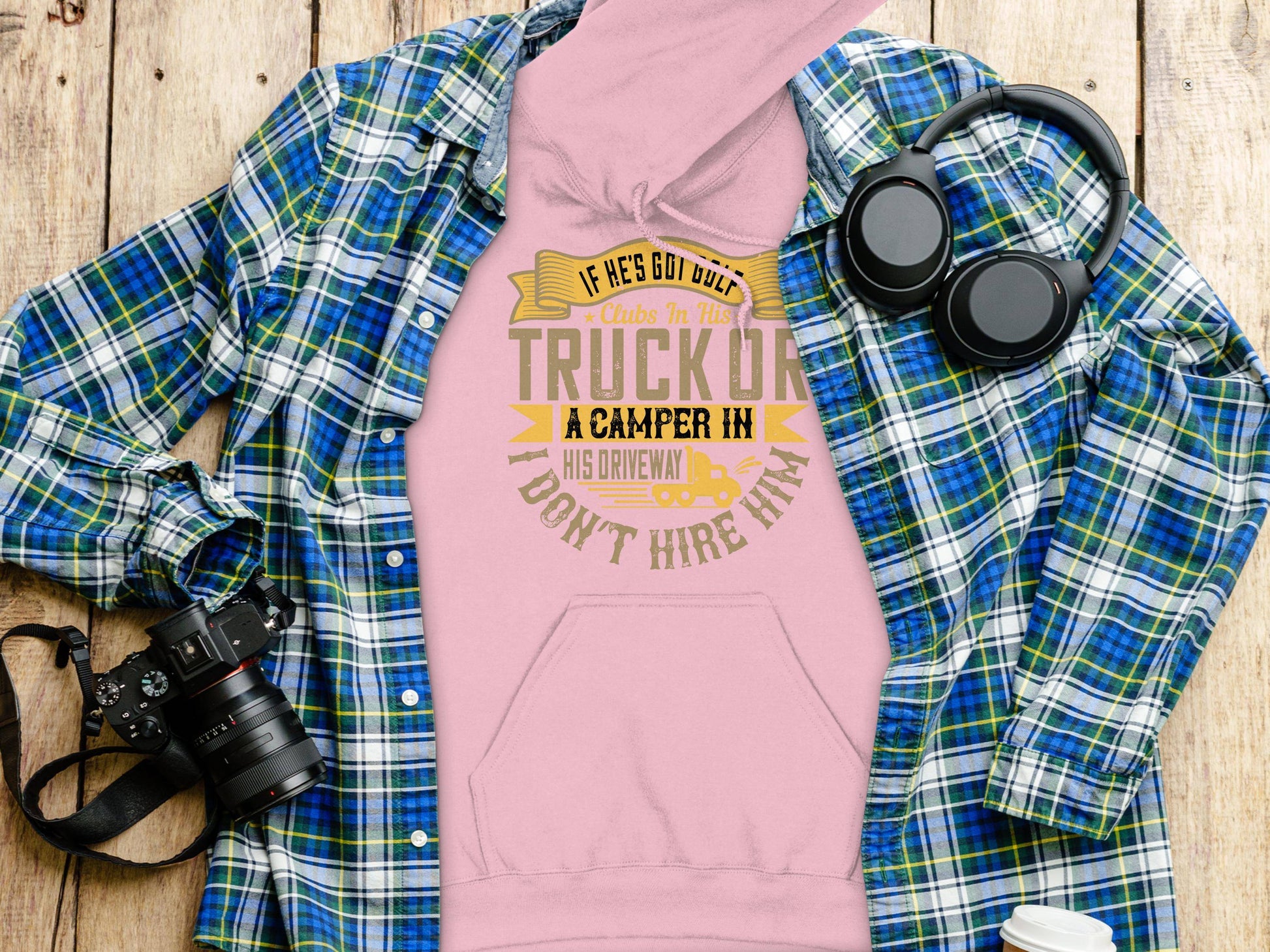 The Garment Graphics pink hoodie, which humorously states If hes got both a truck or a camper in his driveway, dont hire him, rests on a wooden surface. A blue plaid shirt, headphones, a camera, and sunglasses complete the quirky gift display.