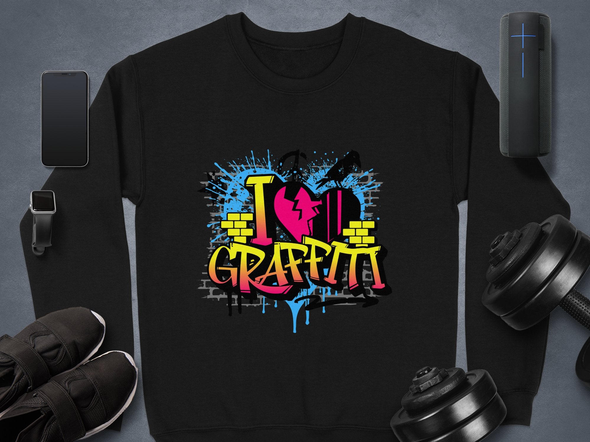 A black Garment Graphics sweatshirt displays vibrant graffiti-style text, I ♥ Graffiti, with heart and brick wall elements. It is surrounded by a smartphone, smartwatch, black sneakers, a Bluetooth speaker, and dumbbells.