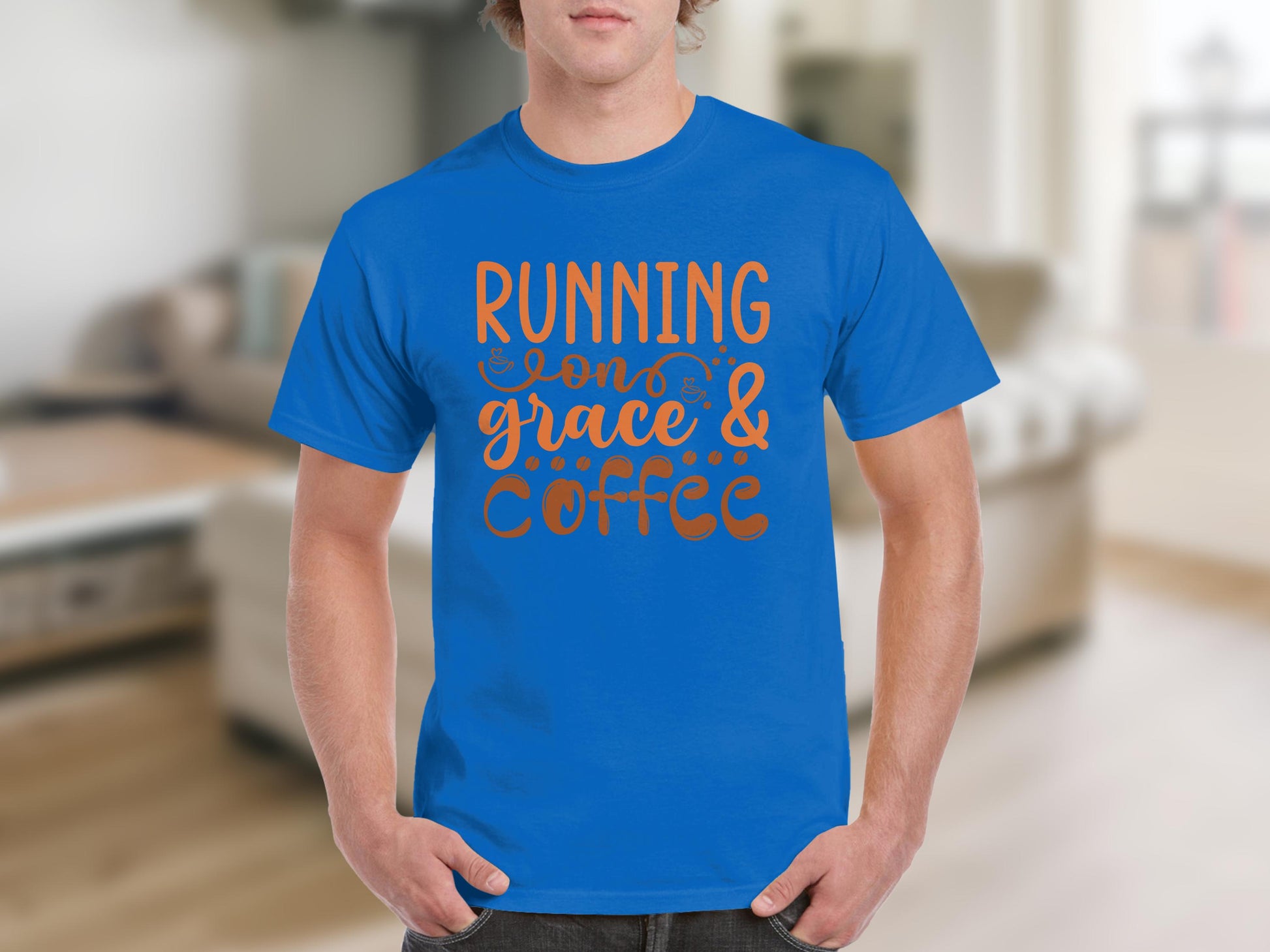 A person indoors wears a blue Garment Graphics t-shirt featuring the bold orange text RUNNING on GRACE & COFFEE, embodying inspirational sayings. The rooms furniture is blurred in the background.