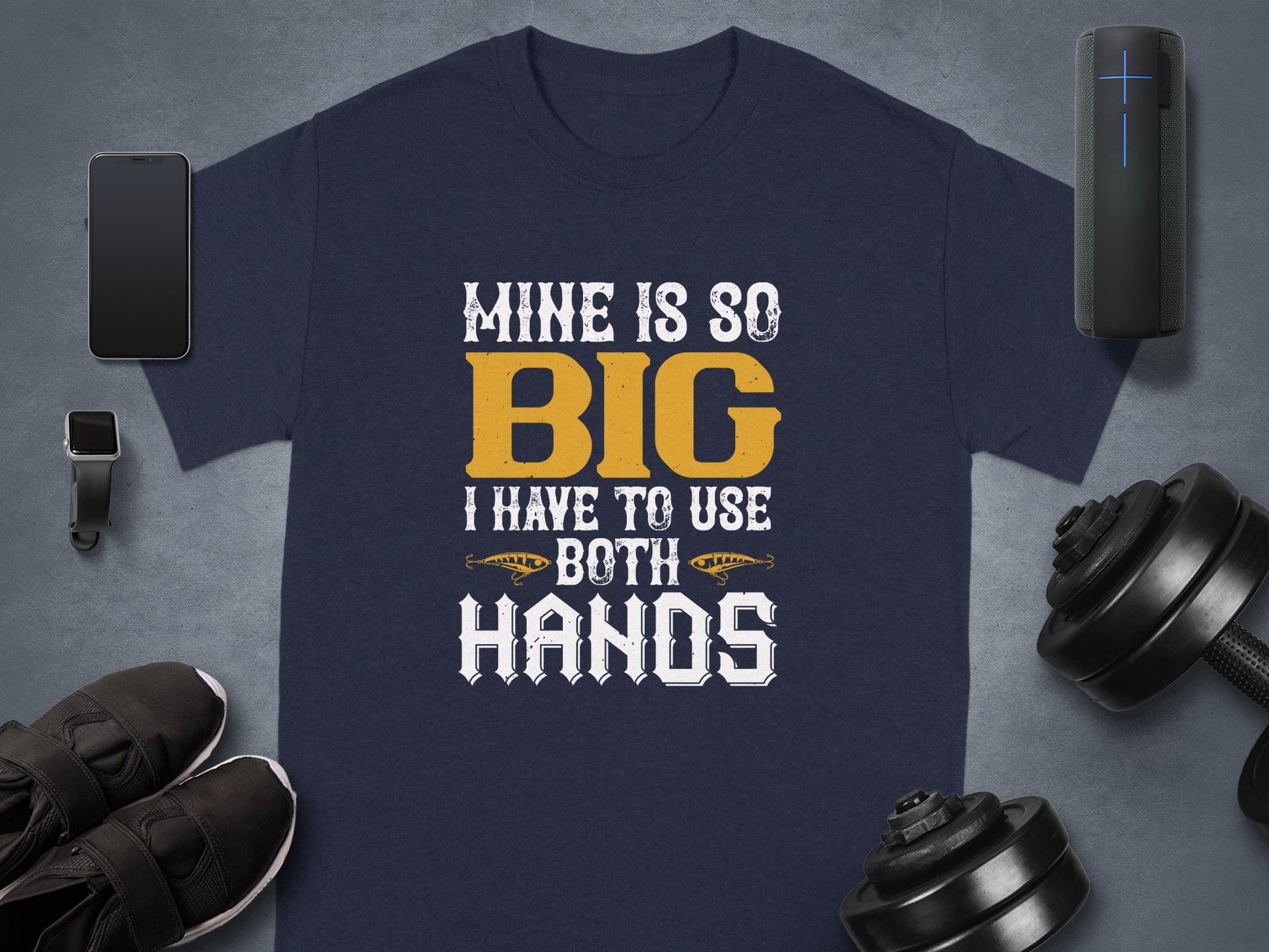 Garment Graphics navy fishing-themed tee features the phrase Mine is so big I have to use both hands in bold white and yellow text. It’s artfully displayed with a smartphone, smartwatch, speaker, dumbbells, shoes, and water bottle on a gray surface—a humorous gift for fishing enthusiasts.