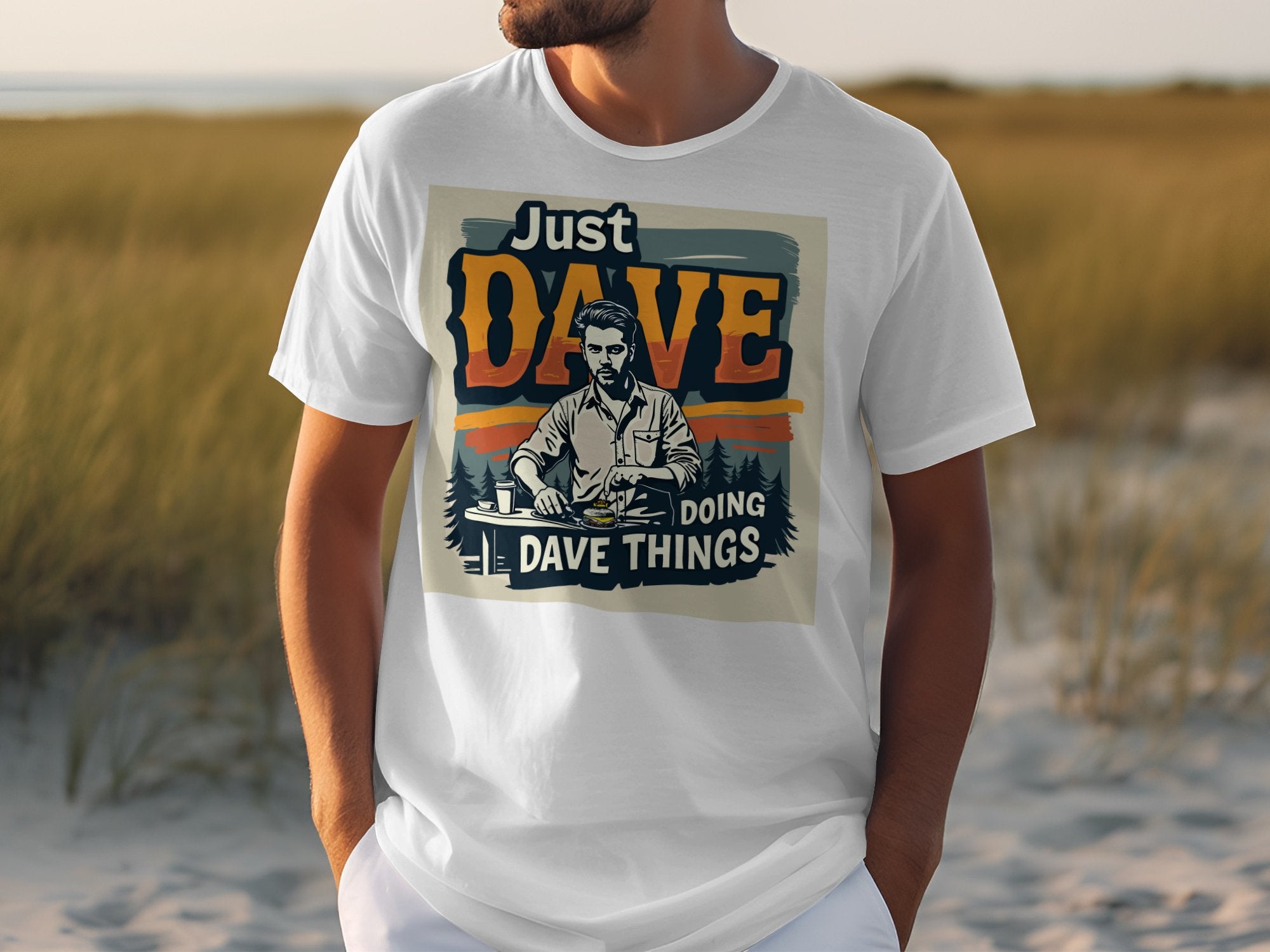 A person is outdoors in a classic Garment Graphics T-shirt saying Just Dave Doing Dave Things, with an illustration of a bearded man holding a drink. The grassy and sandy backdrop enhances the carefree vibe.