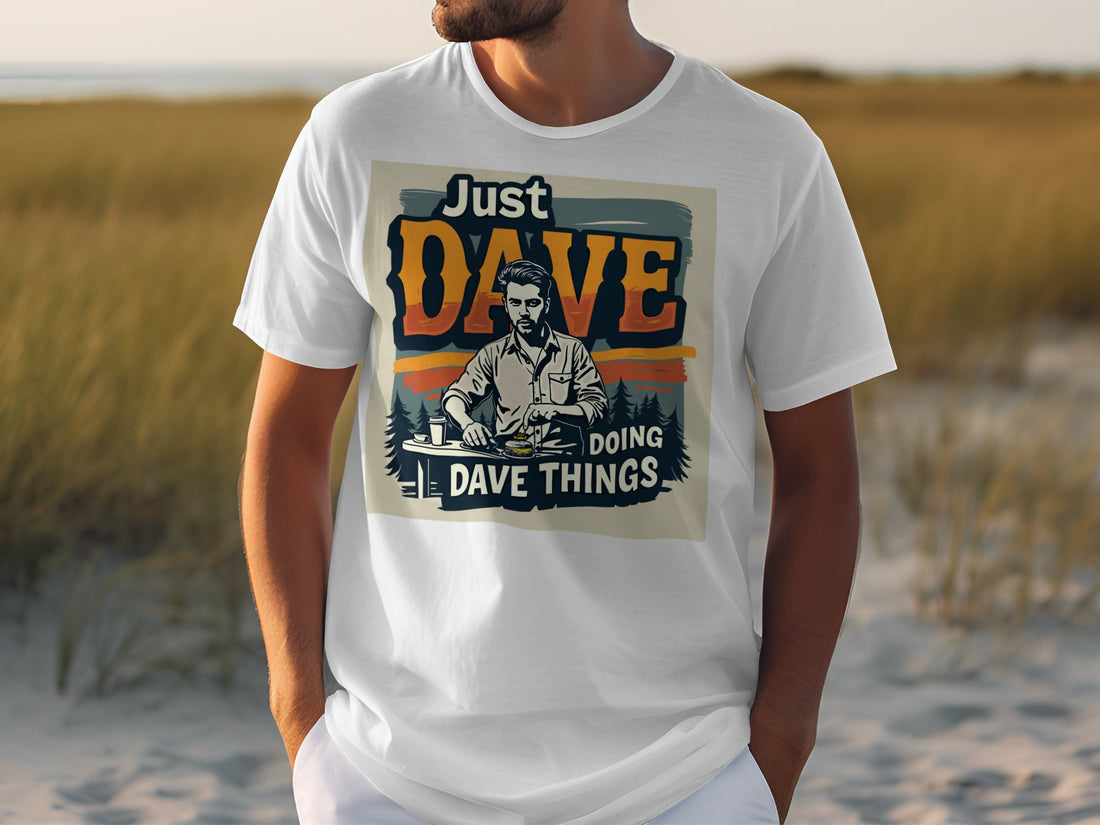 A person is outdoors in a classic Garment Graphics T-shirt saying Just Dave Doing Dave Things, with an illustration of a bearded man holding a drink. The grassy and sandy backdrop enhances the carefree vibe.