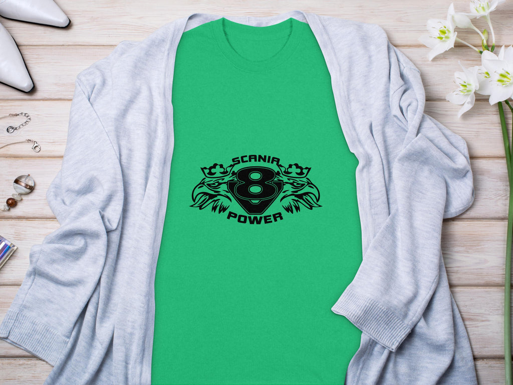 A green T-shirt featuring a black Scania 8 Power logo and two griffins by Garment Graphics lies flat, highlighting sleek design. Its paired with a light gray cardigan on wood, adorned with white orchids and jewelry.