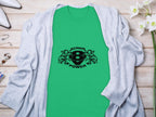A green T-shirt featuring a black Scania 8 Power logo and two griffins by Garment Graphics lies flat, highlighting sleek design. Its paired with a light gray cardigan on wood, adorned with white orchids and jewelry.