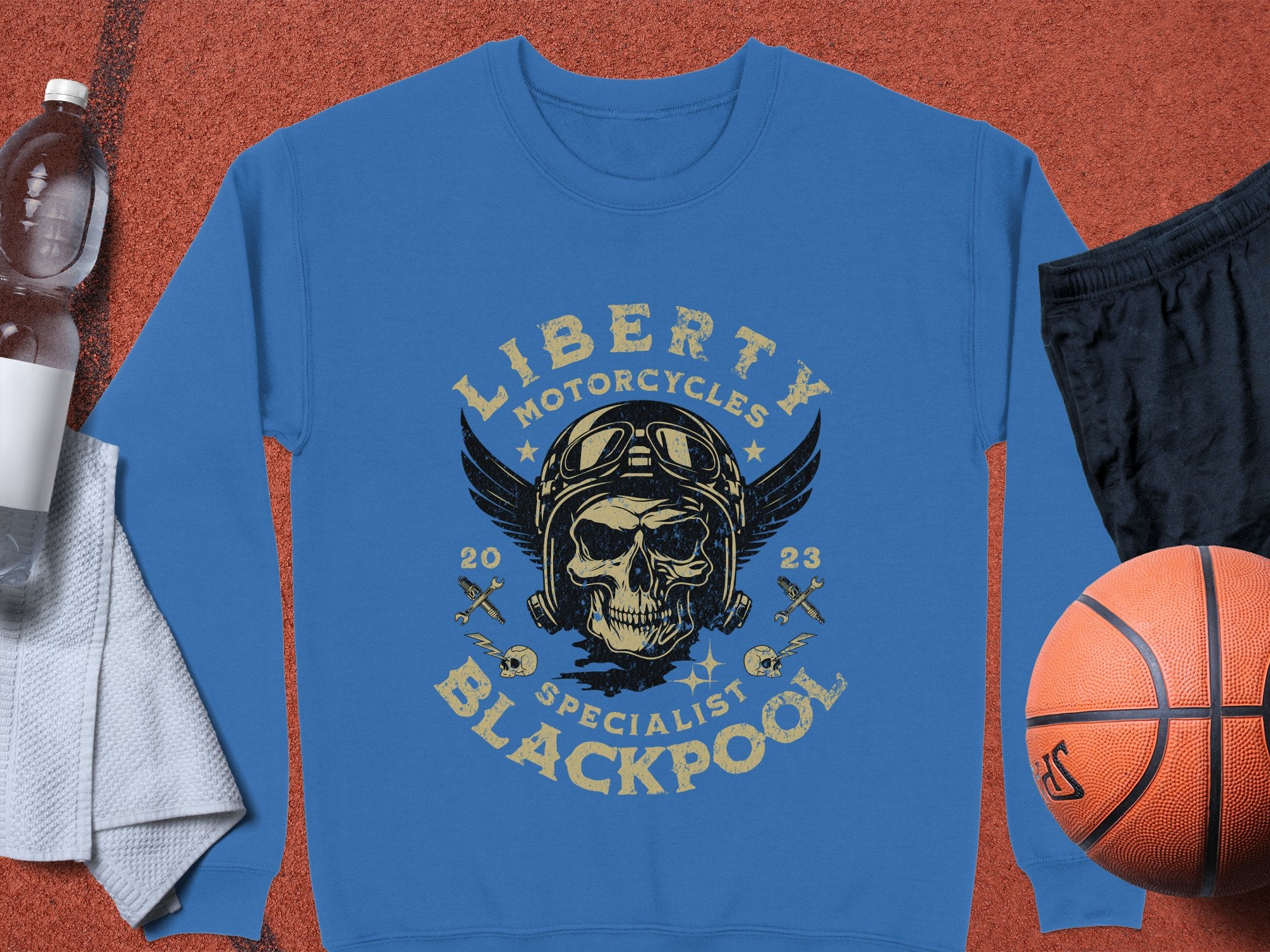 The Liberty Motorcycles Specialist Blackpool 2023 blue sweatshirt by Garment Graphics showcases a skull with goggles and a helmet. Pair it with a white towel, water bottle, black shorts, and an orange basketball for a standout look.