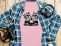 A Garment Graphics pink graphic T-shirt with a black-and-white race car and checkered flag design lies on a wooden surface, surrounded by a blue plaid shirt, headphones, a camera, and a water bottle.