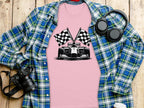 A Garment Graphics pink graphic T-shirt with a black-and-white race car and checkered flag design lies on a wooden surface, surrounded by a blue plaid shirt, headphones, a camera, and a water bottle.