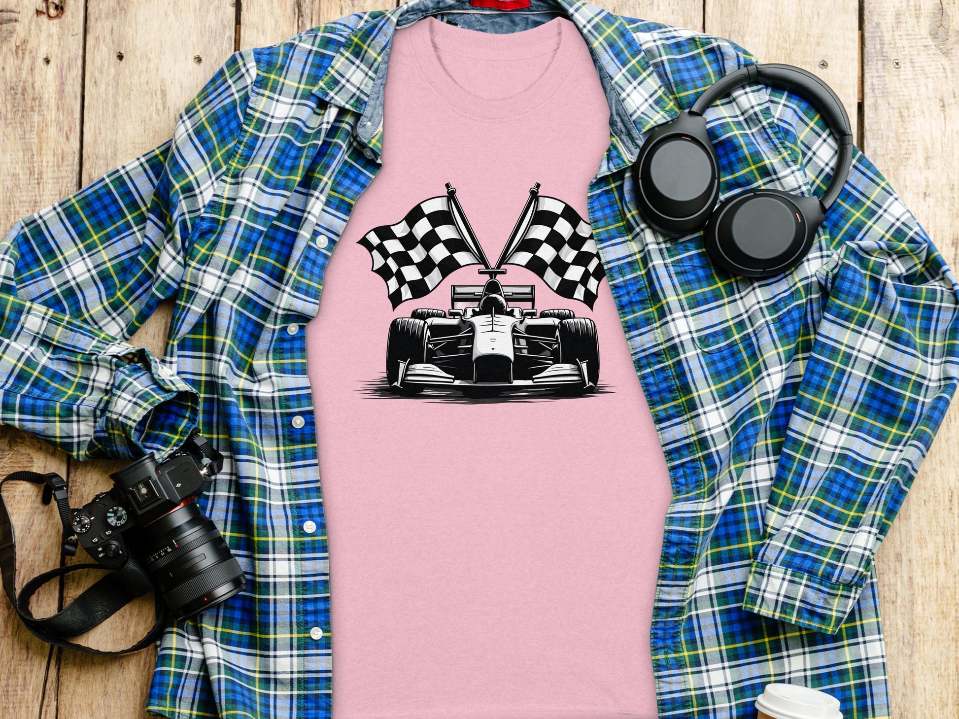 A Garment Graphics pink graphic T-shirt with a black-and-white race car and checkered flag design lies on a wooden surface, surrounded by a blue plaid shirt, headphones, a camera, and a water bottle.