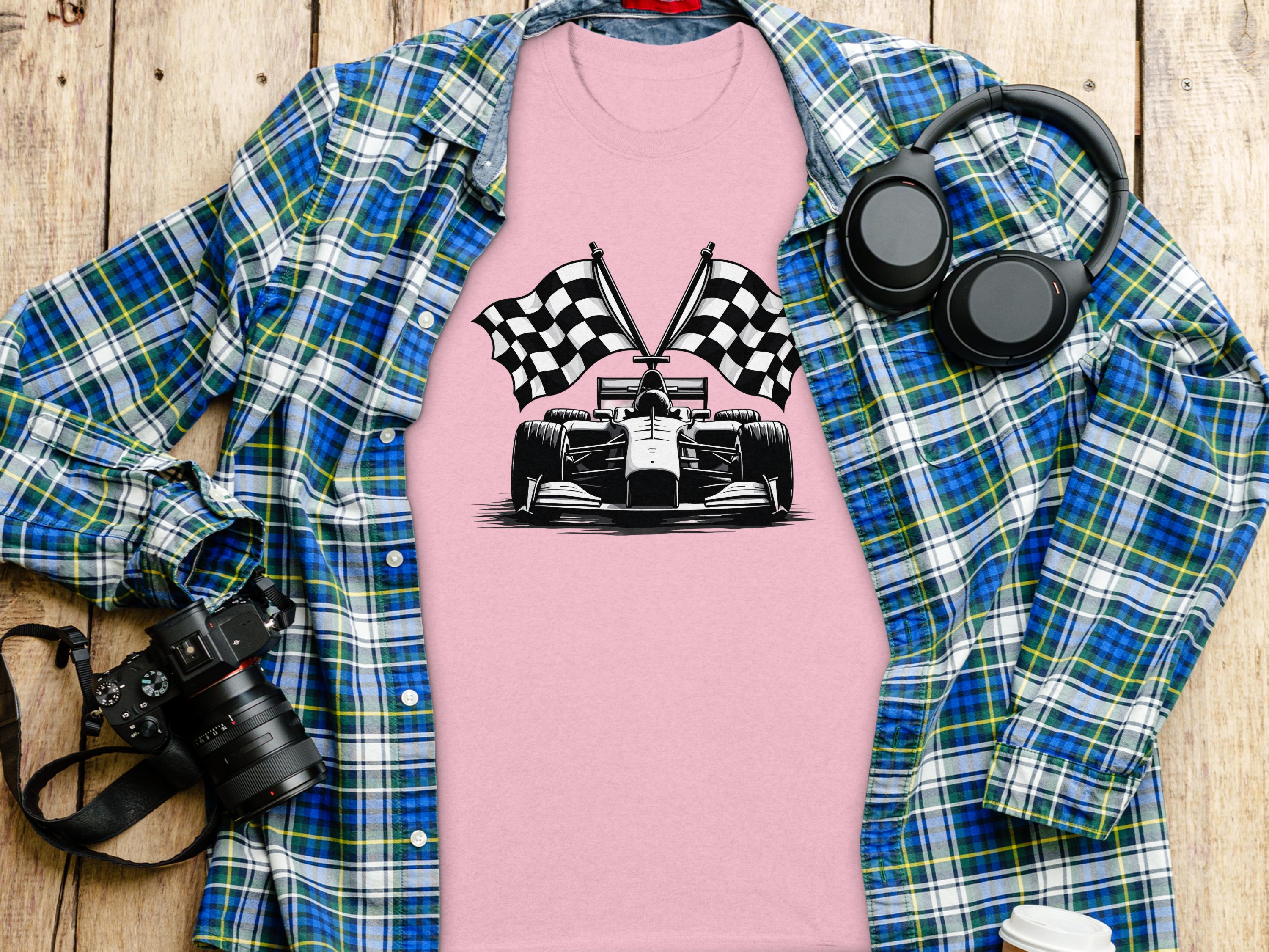 A Garment Graphics pink graphic T-shirt with a black-and-white race car and checkered flag design lies on a wooden surface, surrounded by a blue plaid shirt, headphones, a camera, and a water bottle.