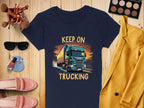 The Garment Graphics Keep on Trucking T-shirt in navy blue, ideal for trucking fans, boasts a striking truck design. Displayed on wood, it pairs with a mustard blazer, makeup brushes, sunglasses, nail polish, and pink loafers.