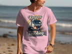 A person in a light pink Garment Graphics T-shirt, showcasing a truck graphic and Keep on Trucking text, stands on the sandy beach with the ocean in view—a great look for truck lovers soaking up seaside vibes.