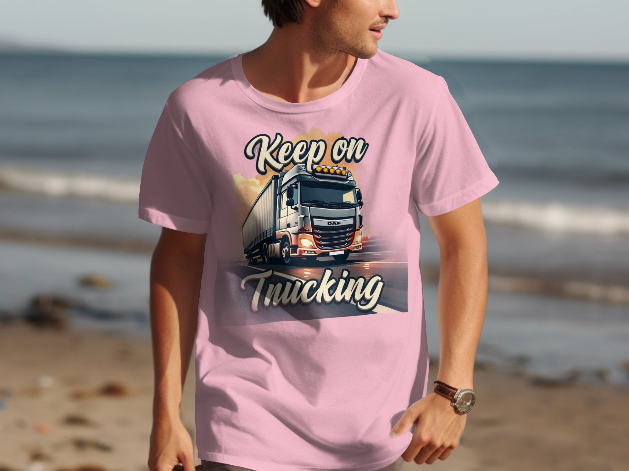 A person in a light pink Garment Graphics T-shirt, showcasing a truck graphic and Keep on Trucking text, stands on the sandy beach with the ocean in view—a great look for truck lovers soaking up seaside vibes.