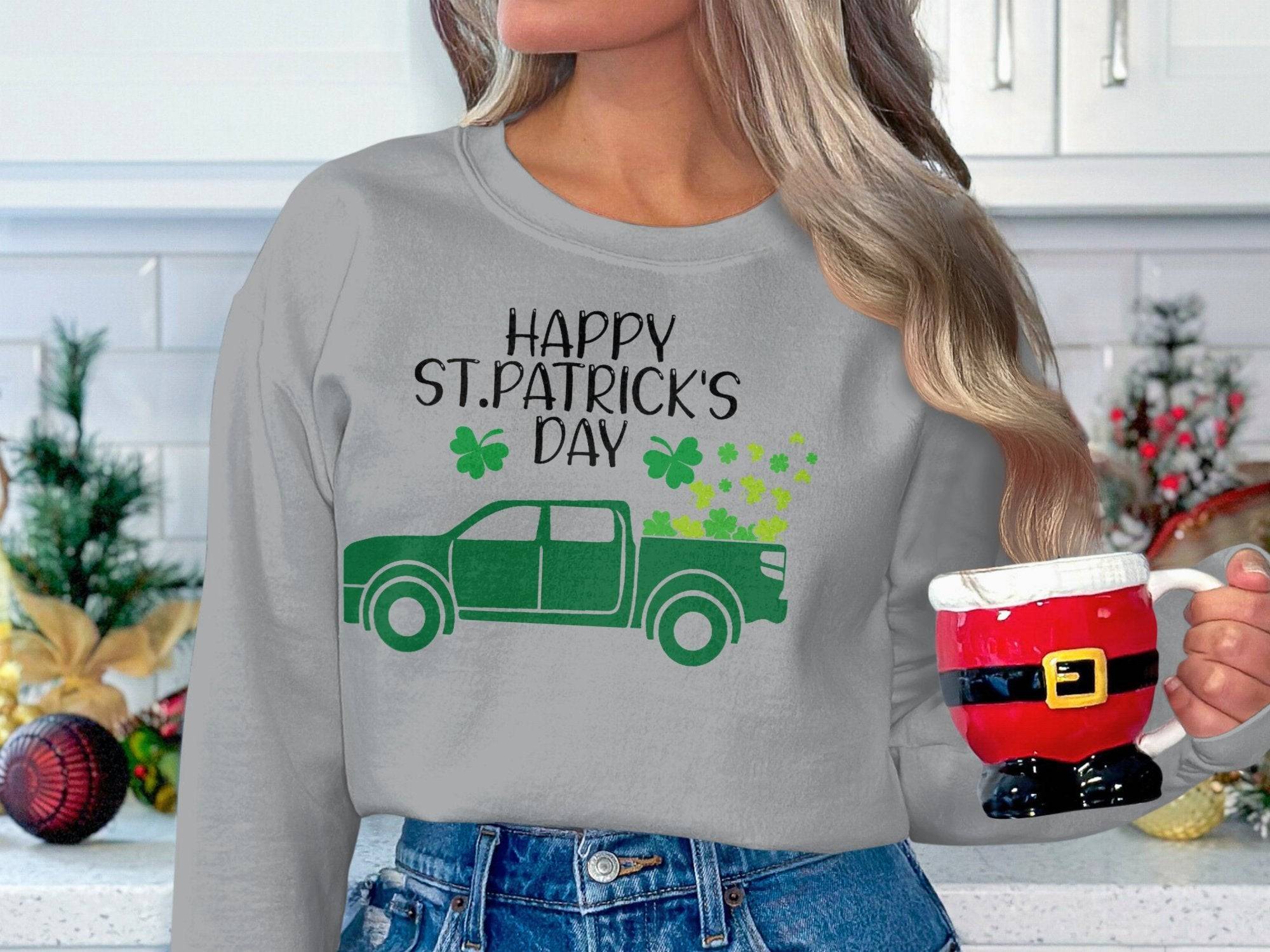 In the kitchen, someone holds a Santa-themed mug while wearing Garment Graphics festive sweatshirt featuring a green truck and shamrocks with Happy St. Patricks Day. The background is filled with holiday decorations.