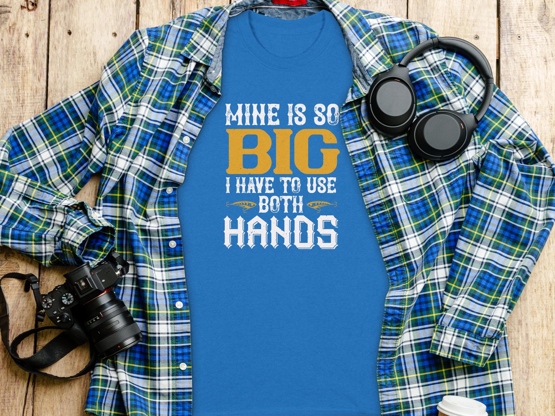 A blue T-shirt from Garment Graphics playfully caters to fishing enthusiasts with the text Mine is so big I have to use both hands. Styled with a plaid shirt, camera, and headphones on a wooden surface, it adds humor to any fishing-themed ensemble.