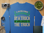 Garment Graphics offers a unisex blue sweatshirt with a vibrant green, yellow, and white design that humorously reads, If an Englishman gets run down by a truck he apologizes to the truck, surrounded by items like a camera, lens, bowl of kiwi, map, and book.