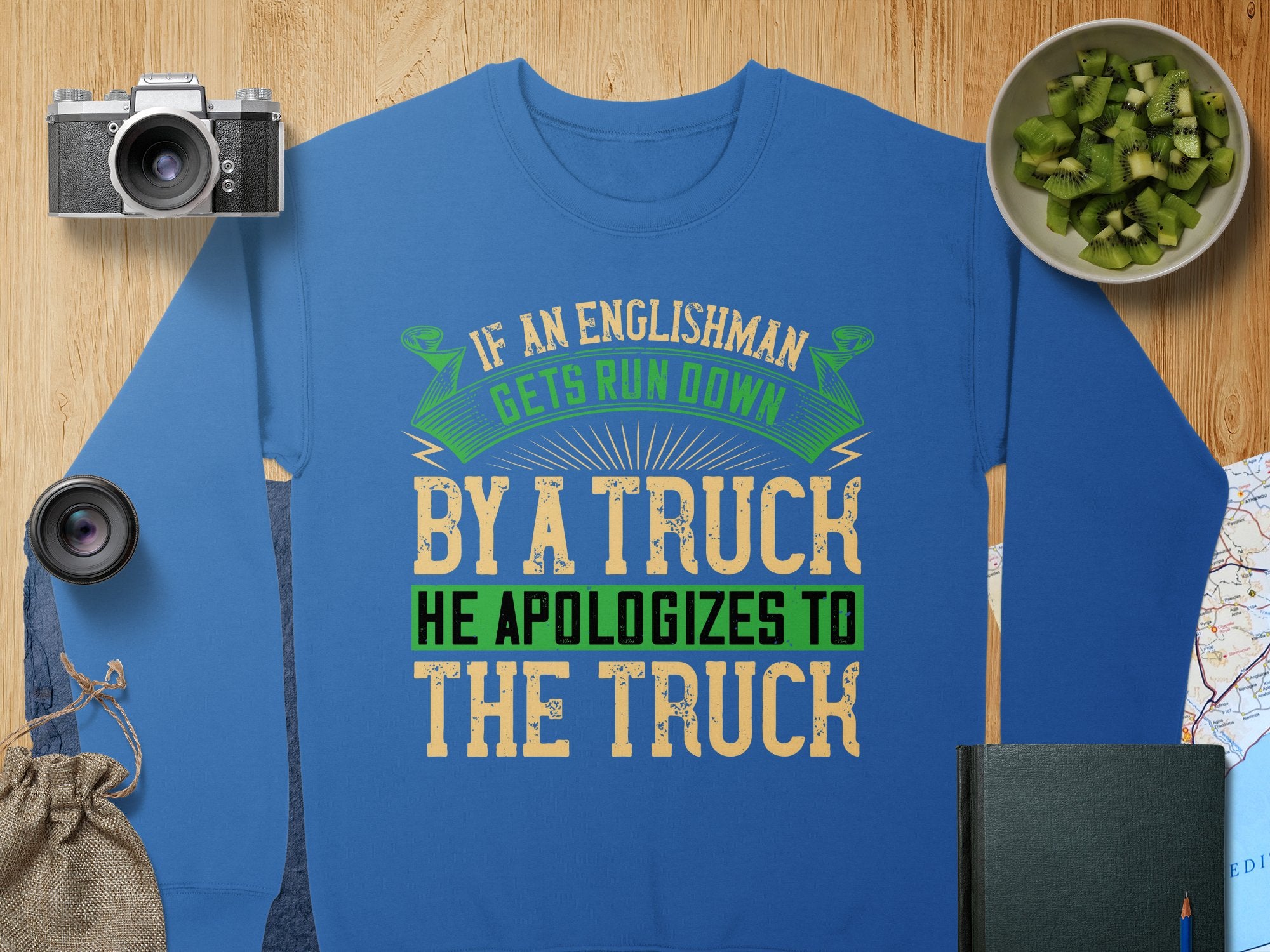 Garment Graphics offers a unisex blue sweatshirt with a vibrant green, yellow, and white design that humorously reads, If an Englishman gets run down by a truck he apologizes to the truck, surrounded by items like a camera, lens, bowl of kiwi, map, and book.