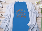 Garment Graphics classic blue T-shirt features This day is powered by coffee in decorative brown lettering. Styled flat with a light gray cardigan, white shoes, jewelry, and white flowers on a wooden surface, it embodies a positive vibe.