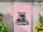 Garment Graphics pink T-shirt with a bold truck graphic and Keep on Trucking text is displayed flat on concrete, surrounded by green foliage and vibrant yellow and red flowers.