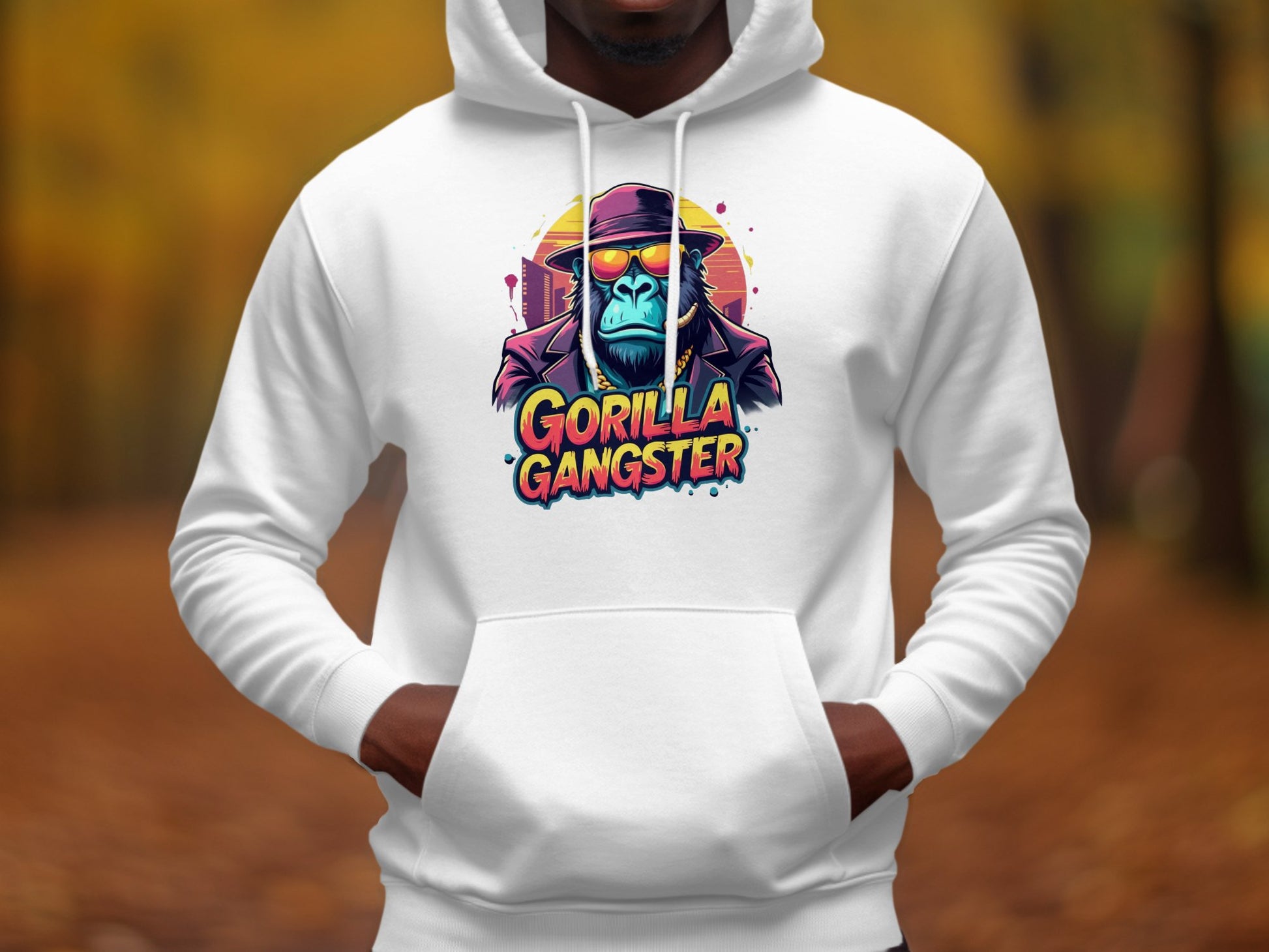 A person wears a Garment Graphics white streetwear hoodie featuring a graphic of a blue gorilla in a hat and sunglasses, with the bold text Gorilla Gangster. Set amid blurry autumn leaves outdoors, it captures the edgy spirit of gangster hoodies.