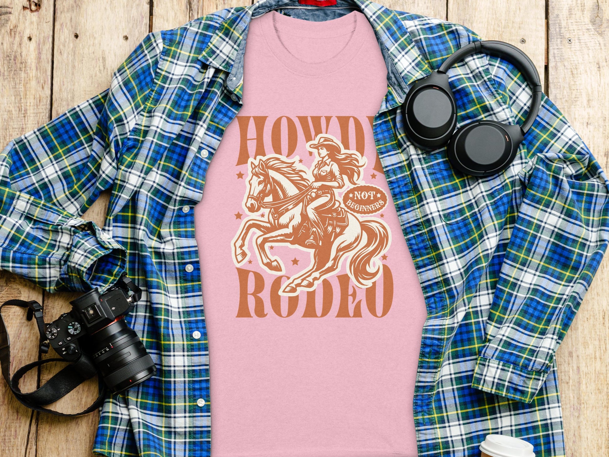 A Garment Graphics pink Howdy Rodeo t-shirt, featuring a cowgirl on horseback, is stylishly layered over a green and blue plaid shirt on wood, paired with essentials like a camera, headphones, and water bottle—a must-have rodeo tee for enthusiasts.