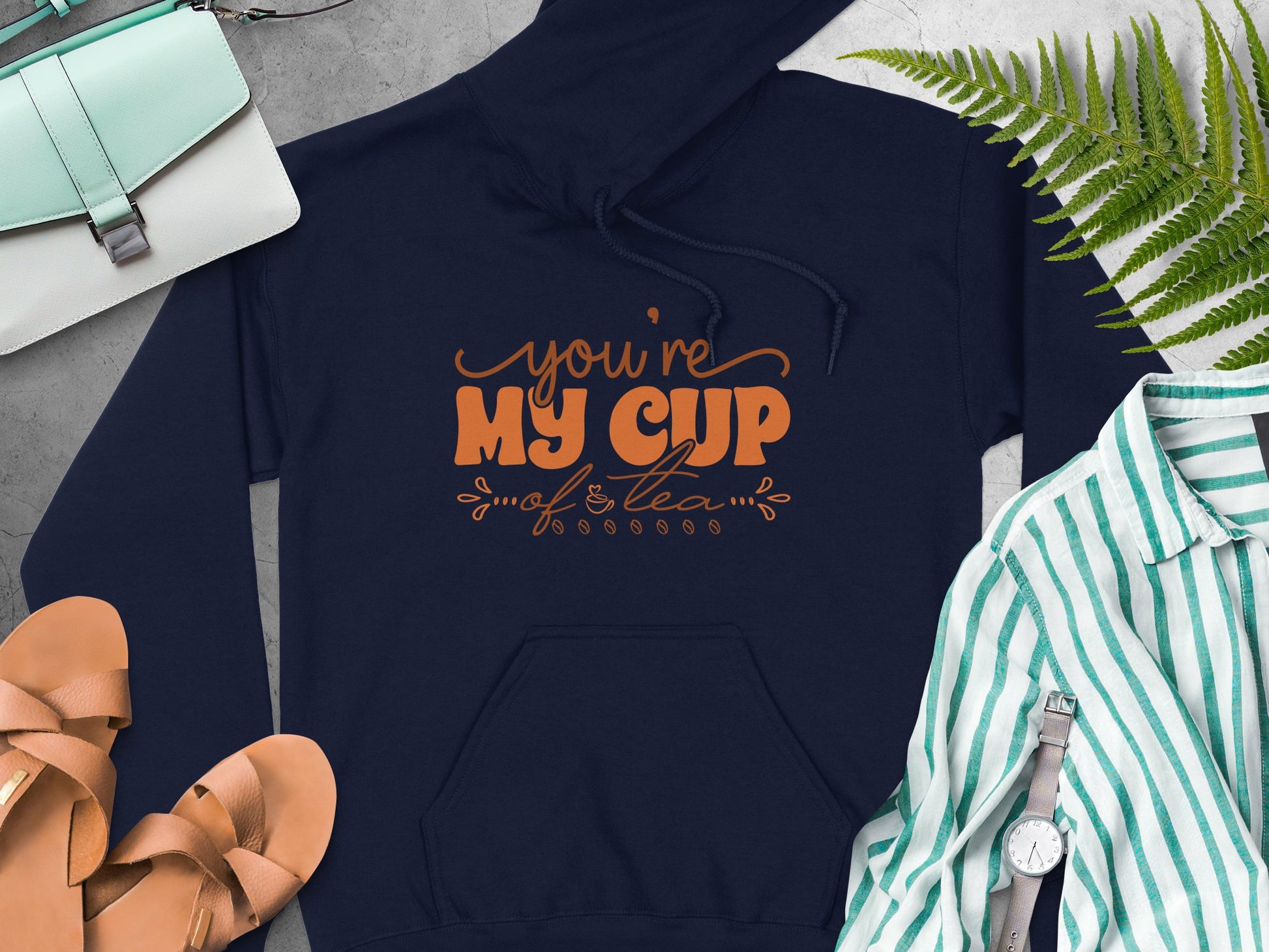 A navy blue hoodie with Youre my cup of tea in orange by Garment Graphics lies beside a white and green striped shirt, brown sandals, a wristwatch, a fern leaf, and a white handbag with a green strap—perfect for cozy moments.