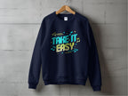 A black sweatshirt hangs on a wooden hanger against a gray concrete wall. Designed by Garment Graphics, it features Take It Easy in colorful letters with musical notes and arrows. Unisex sizing makes it ideal for sharing your laid-back style.
