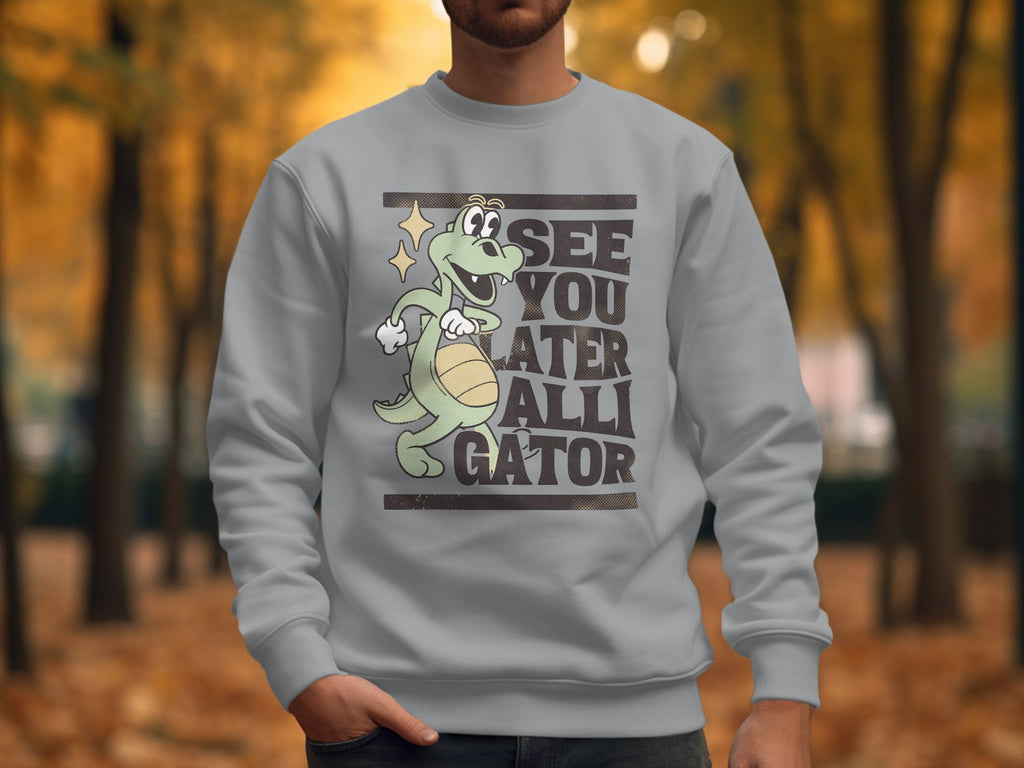 Wearing a Garment Graphics sweatshirt featuring a cartoon alligator with a ball and See You Later Alligator text, the person enjoys comfort in unisex sizing. Blurred autumn leaves create a scenic park backdrop.