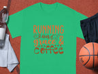 The Garment Graphics green T-shirt displays the phrase Running on Grace & Coffee in whimsical orange and brown font. Accompanying it are a basketball, black shorts, white towel, and a plastic water bottle on a textured orange background.