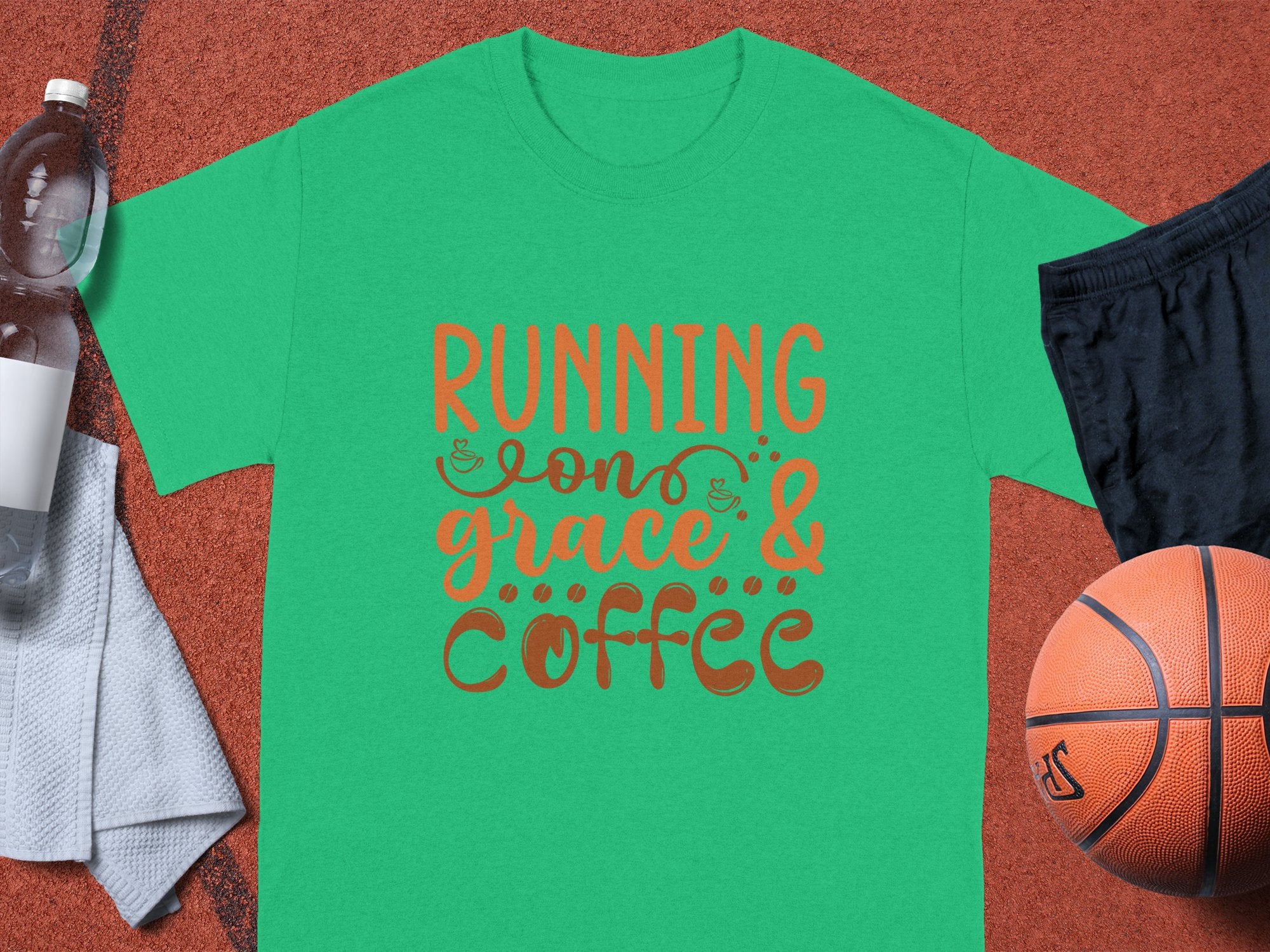 The Garment Graphics green T-shirt displays the phrase Running on Grace & Coffee in whimsical orange and brown font. Accompanying it are a basketball, black shorts, white towel, and a plastic water bottle on a textured orange background.