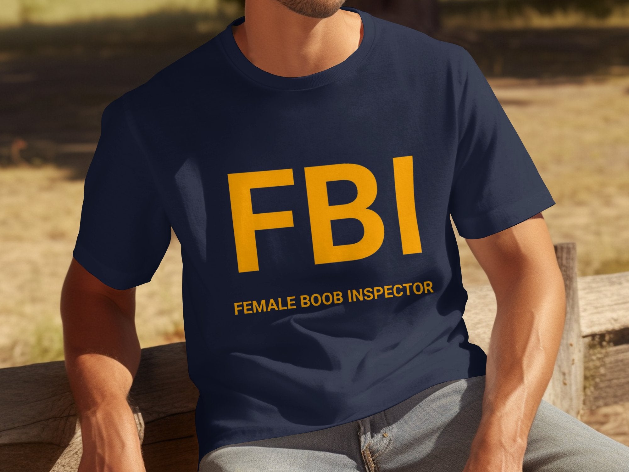 A person sits on a wooden bench wearing a Garment Graphics T-shirt featuring large yellow FBI letters and smaller text reading Female Boob Inspector. This humorous design pops against the grassy backdrop.