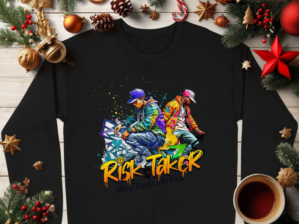 A black sweatshirt from Garment Graphics, showcasing vibrant street dance graphics of two figures and the text “Risk Taker Street Dance,” is placed on a wooden surface amid pine branches, star anise, pine cones, ornaments, and a cozy cup of coffee.