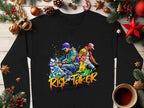 A black sweatshirt from Garment Graphics, showcasing vibrant street dance graphics of two figures and the text “Risk Taker Street Dance,” is placed on a wooden surface amid pine branches, star anise, pine cones, ornaments, and a cozy cup of coffee.