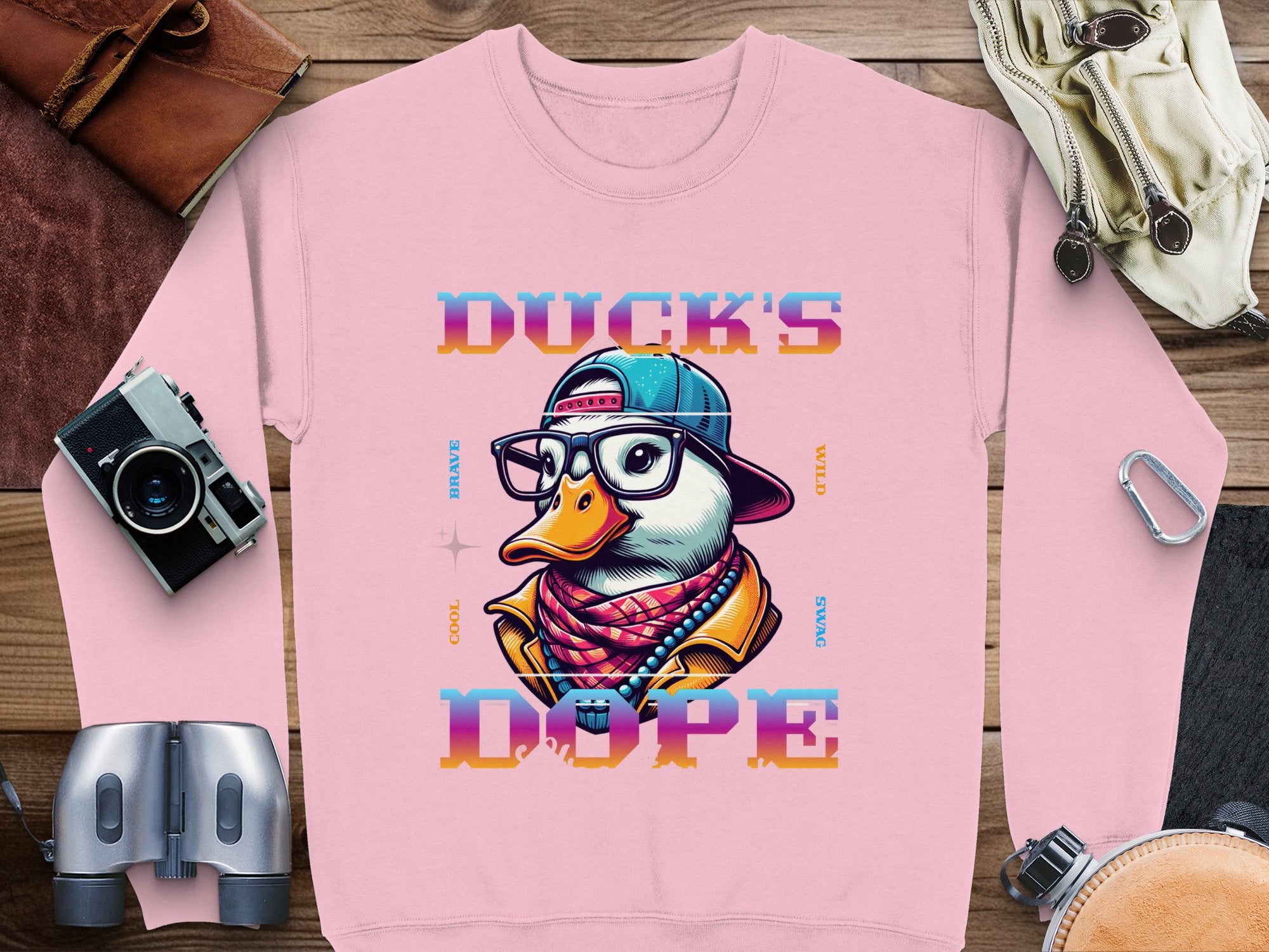 This stylish pink cartoon graphic sweatshirt by Garment Graphics features a duck wearing glasses, a cap, and a vibrant jacket with the bold text DUCKS DOPE. When laid flat, its complemented by travel essentials: a camera, binoculars, backpack, and leather pouch.