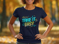 A person wears a navy blue Garment Graphics T-shirt featuring the stylish Take It Easy design with vivid arrows, musical notes, and stars. The background highlights a blurred view of trees in fall foliage.