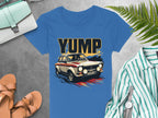 A bold graphic vintage car with YUMP is featured on this blue t-shirt by Garment Graphics. It is displayed on a gray background, accompanied by a striped shirt, brown sandals, light blue handbag, watch, and green fern leaves.
