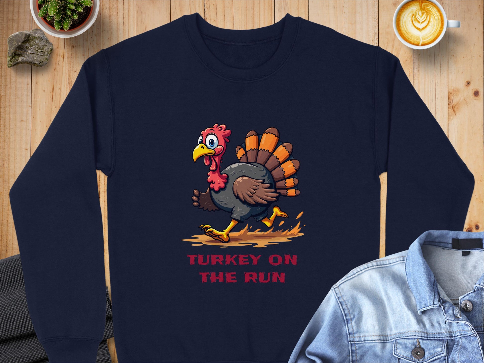 On a wooden surface lies a navy sweatshirt from Garment Graphics featuring a funny cartoon turkey with Turkey on the Run in red, next to a folded denim shirt, latte art coffee cup, and potted succulent—stylish and humorous.