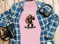 A Garment Graphics pink T-shirt with a Bigfoot design and Admit it, life would be boring without me is placed over a plaid button-up shirt. Surrounding this are headphones, a camera, a coffee cup, and a plant, artfully arranged on wood.