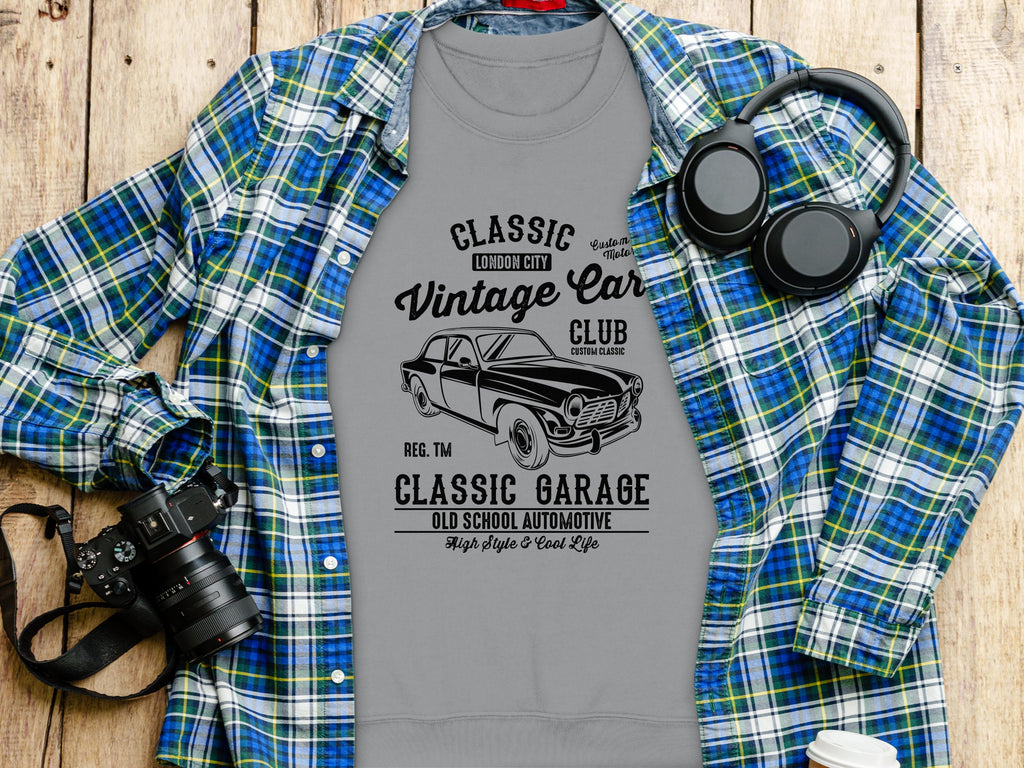 A gray Garment Graphics T-shirt with a vintage car graphic and the text Classic London City Vintage Car Club, Classic Garage, Old School Automotive, High Style & Cool Life is draped over a blue and green plaid shirt. Nearby sit a camera and headphones on the wooden surface.