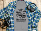 A gray Garment Graphics T-shirt with a vintage car graphic and the text Classic London City Vintage Car Club, Classic Garage, Old School Automotive, High Style & Cool Life is draped over a blue and green plaid shirt. Nearby sit a camera and headphones on the wooden surface.