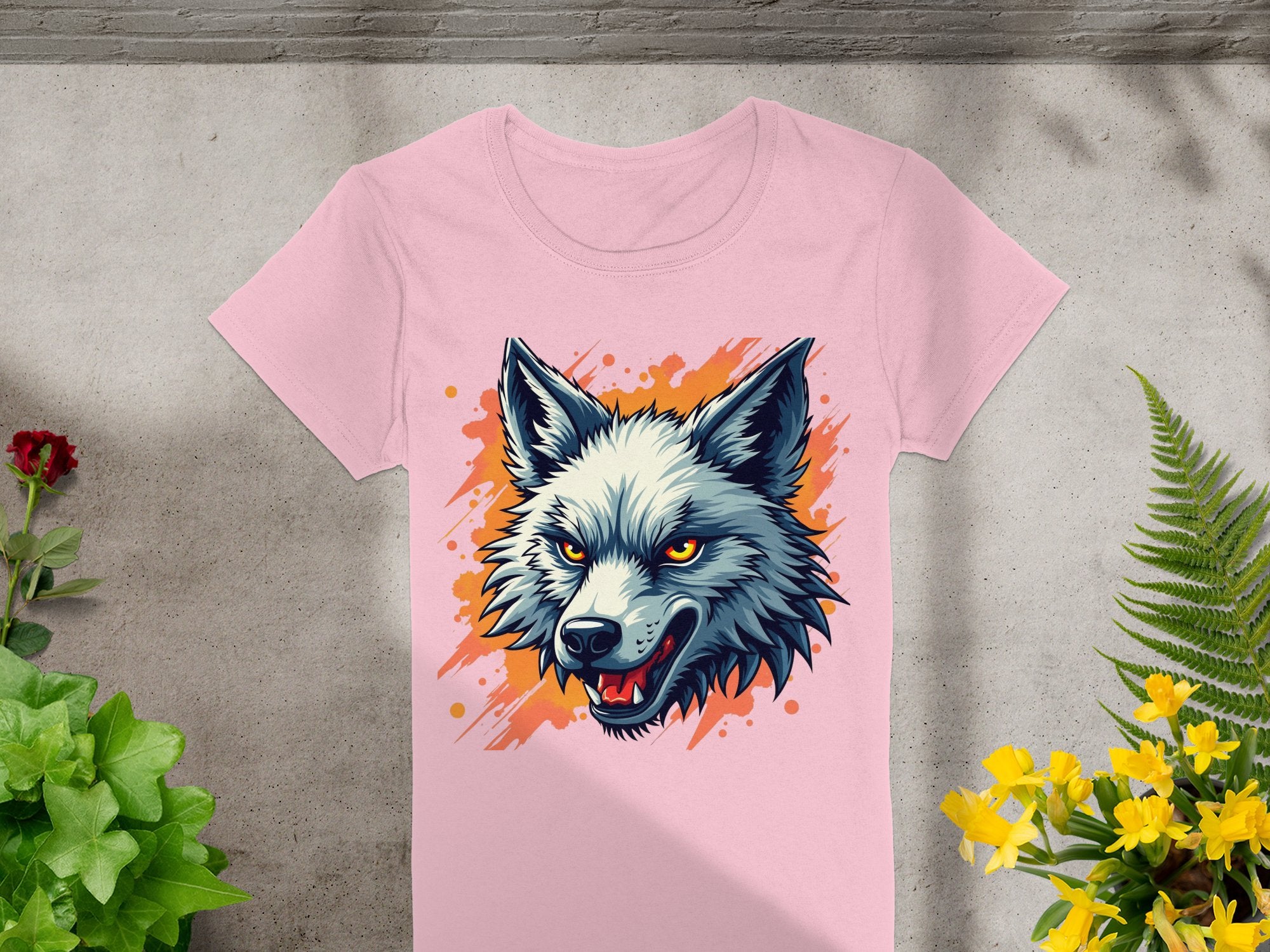 Garment Graphics presents a 100% cotton pink T-shirt featuring a wolfs head with gray and white fur and yellow eyes on an orange splash. Its displayed on a gray surface amidst green and yellow plants—ideal for those with daring personalities.