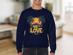A person indoors models Garment Graphics navy blue sweatshirt, highlighting the One Love Sunset design with a tropical sunset, palm trees, an orange sky, a plane overhead, and ONE LOVE in bold yellow and orange letters.