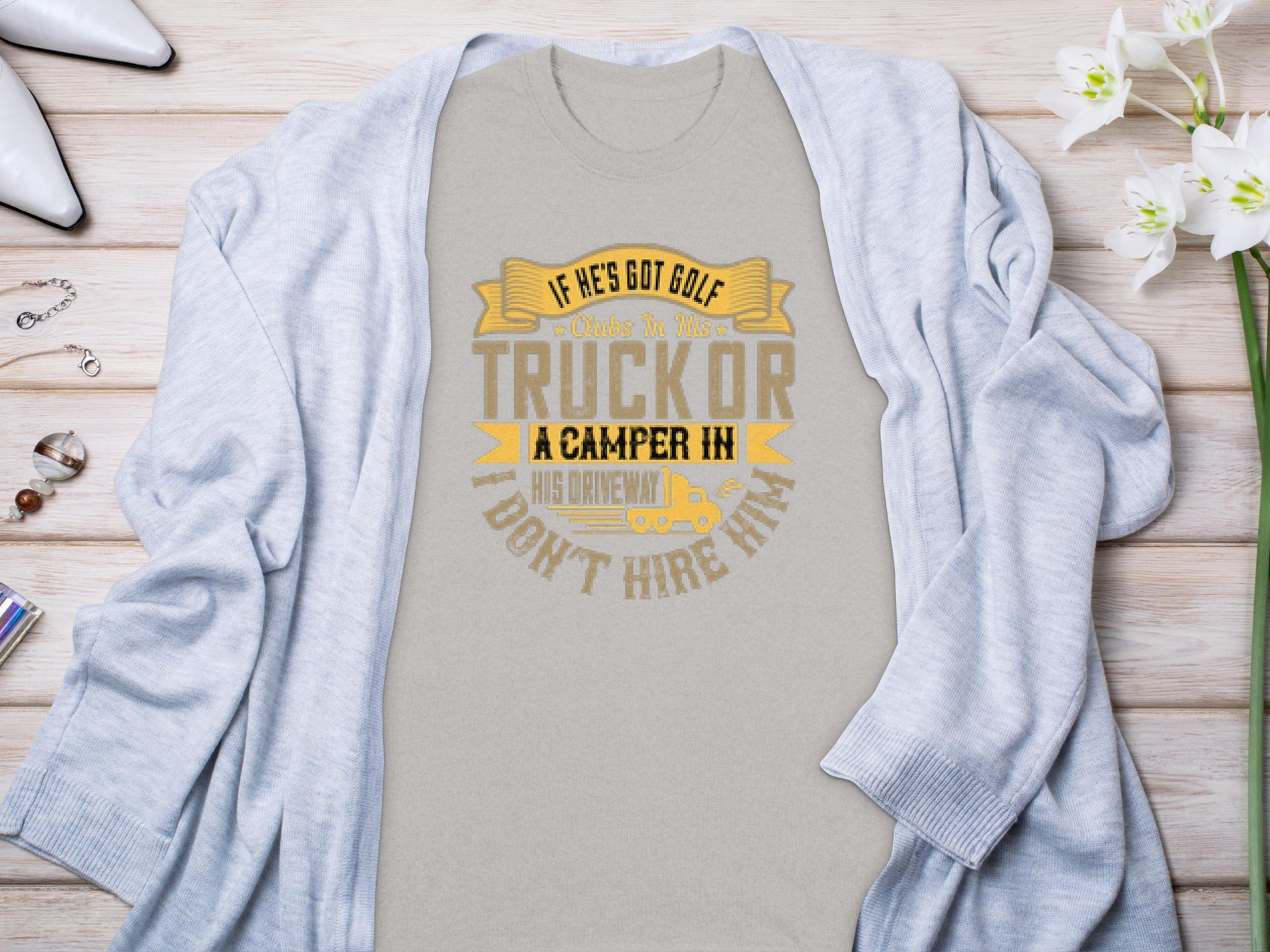 Light gray Garment Graphics t-shirt with the text If hes got golf clubs in his truck or a camper in his driveway, dont hire him in yellow and gray. Ideal for camping or golf fans, styled with a cardigan and surrounded by white flowers and jewelry.