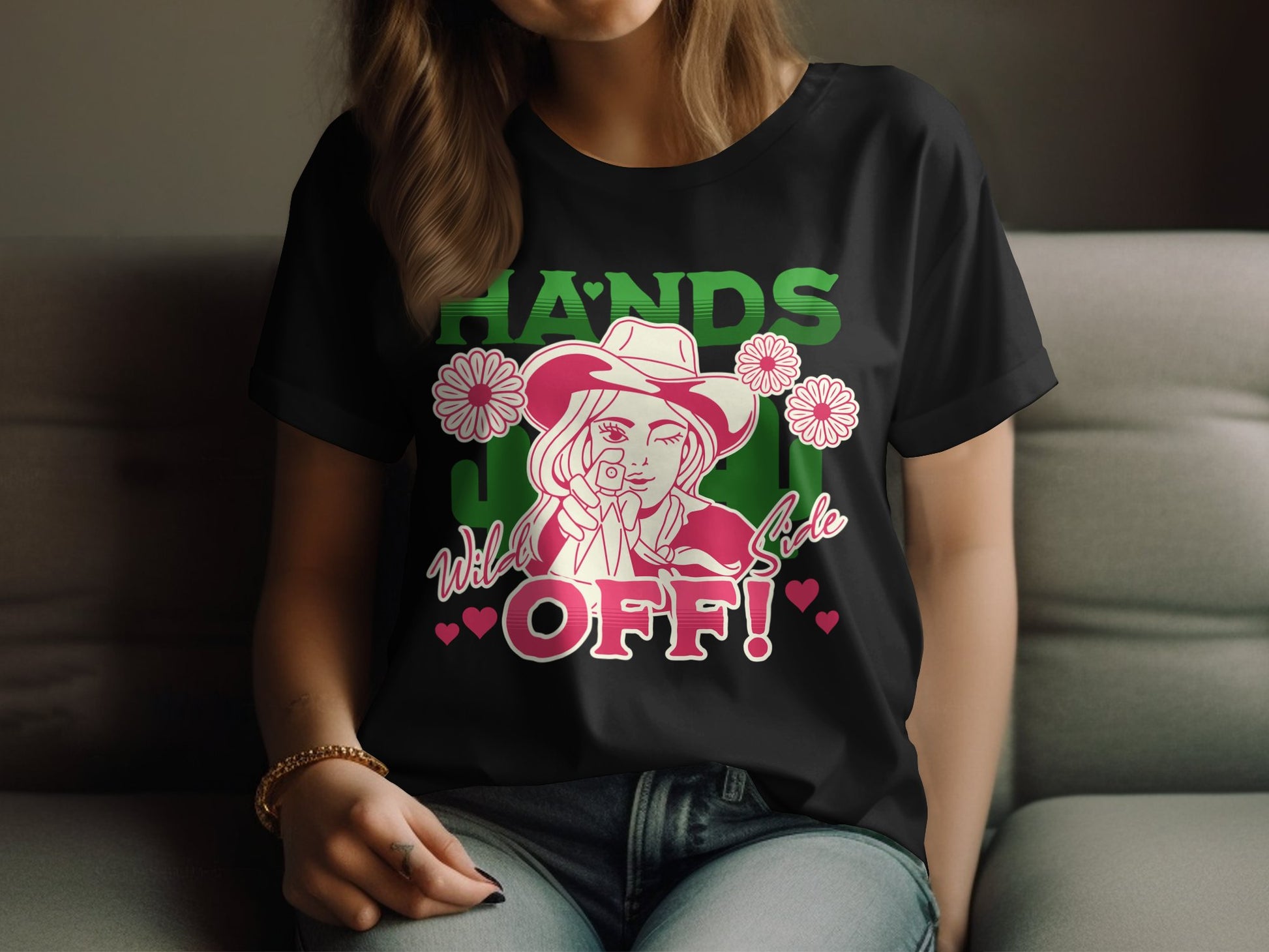 A woman relaxes on a couch wearing a Garment Graphics black T-shirt featuring a bold design of a cowgirl with HANDS OFF! in green letters, accented by pink flowers and hearts, reflecting her unique style.