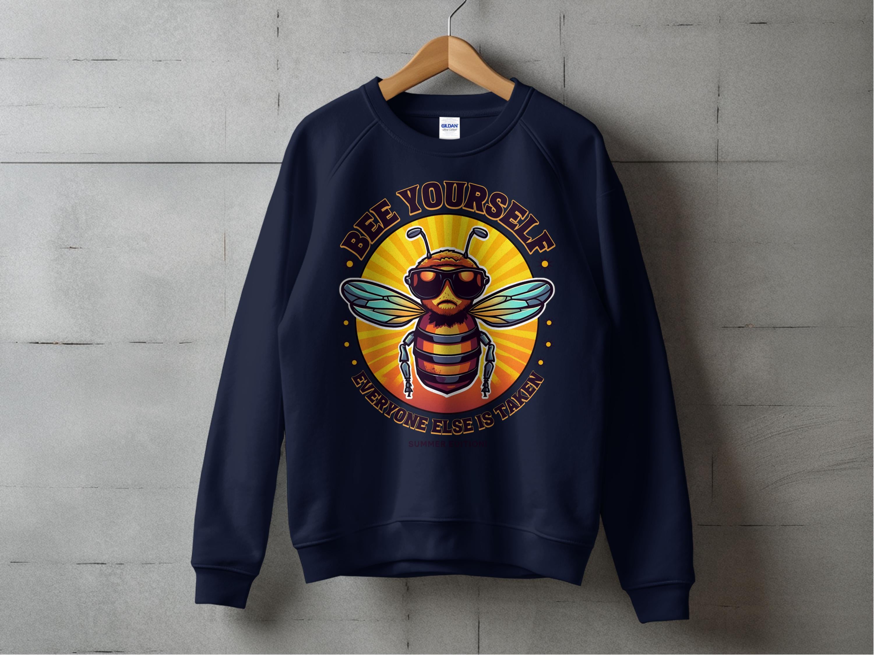 A navy sweatshirt from Garment Graphics exclusive Summer Edition hangs against a concrete wall, featuring a bee in sunglasses and the phrase Bee Yourself, Everyone Else is Taken. Its unisex sizing makes it perfect for everyone!.