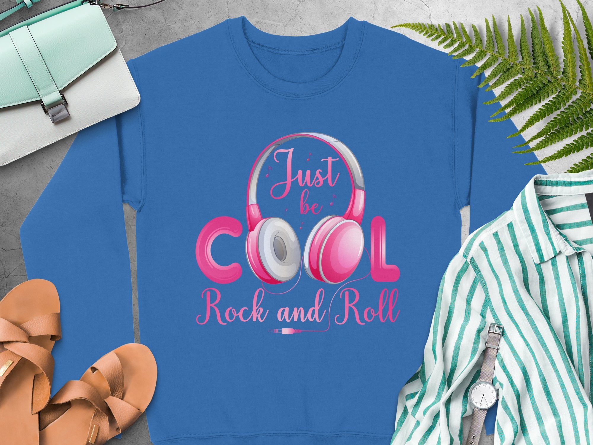 Garment Graphics blue sweatshirt, adorned with Just be Cool, Rock and Roll and headphones, is styled alongside tan sandals, a mint handbag, a striped shirt, a watch, and a fern on a gray surface—ideal for fashion lovers and music enthusiasts.