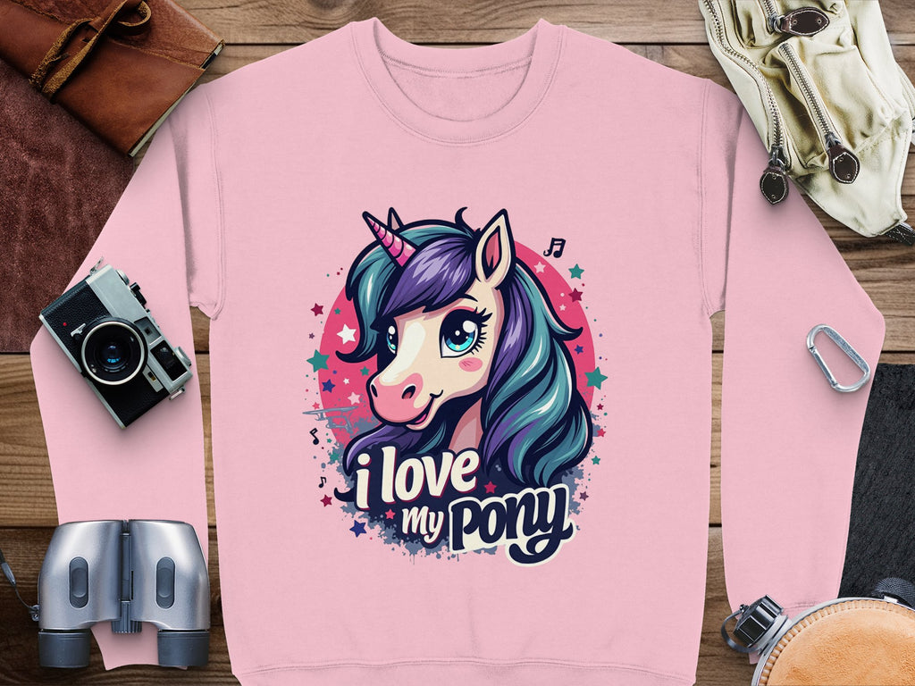 The Garment Graphics pink sweatshirt showcases a cartoon unicorn with a colorful mane and horn, surrounded by stars and music notes, with I love my pony text. Ideal for graphic sweatshirt fans, its styled flat beside a camera, binoculars, and other accessories.