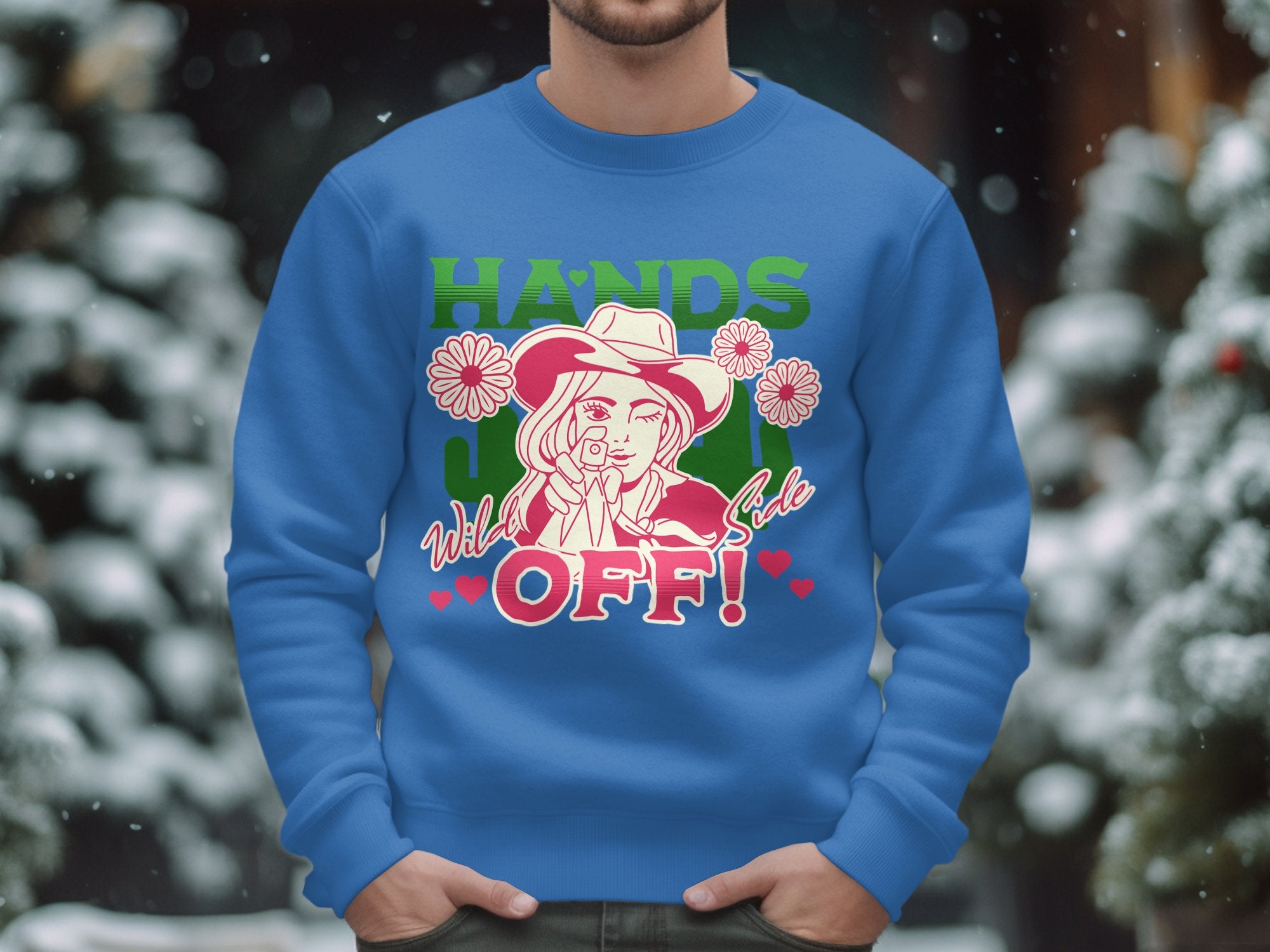 A man sports a blue Garment Graphics sweatshirt from the Wild Side Sweatshirts collection, showcasing bold graphics of a woman in a hat with daisies and the words Hands Off! Unisex sizing ensures comfort for all, set against a background of snowy trees.
