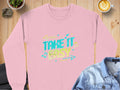 A pink Garment Graphics sweatshirt with Take It Easy in colorful retro-style text sits on a wooden table, surrounded by a latte, denim jacket, plant, and rock. The unisex fit and vibrant design make it ideal for anyone seeking style and comfort.