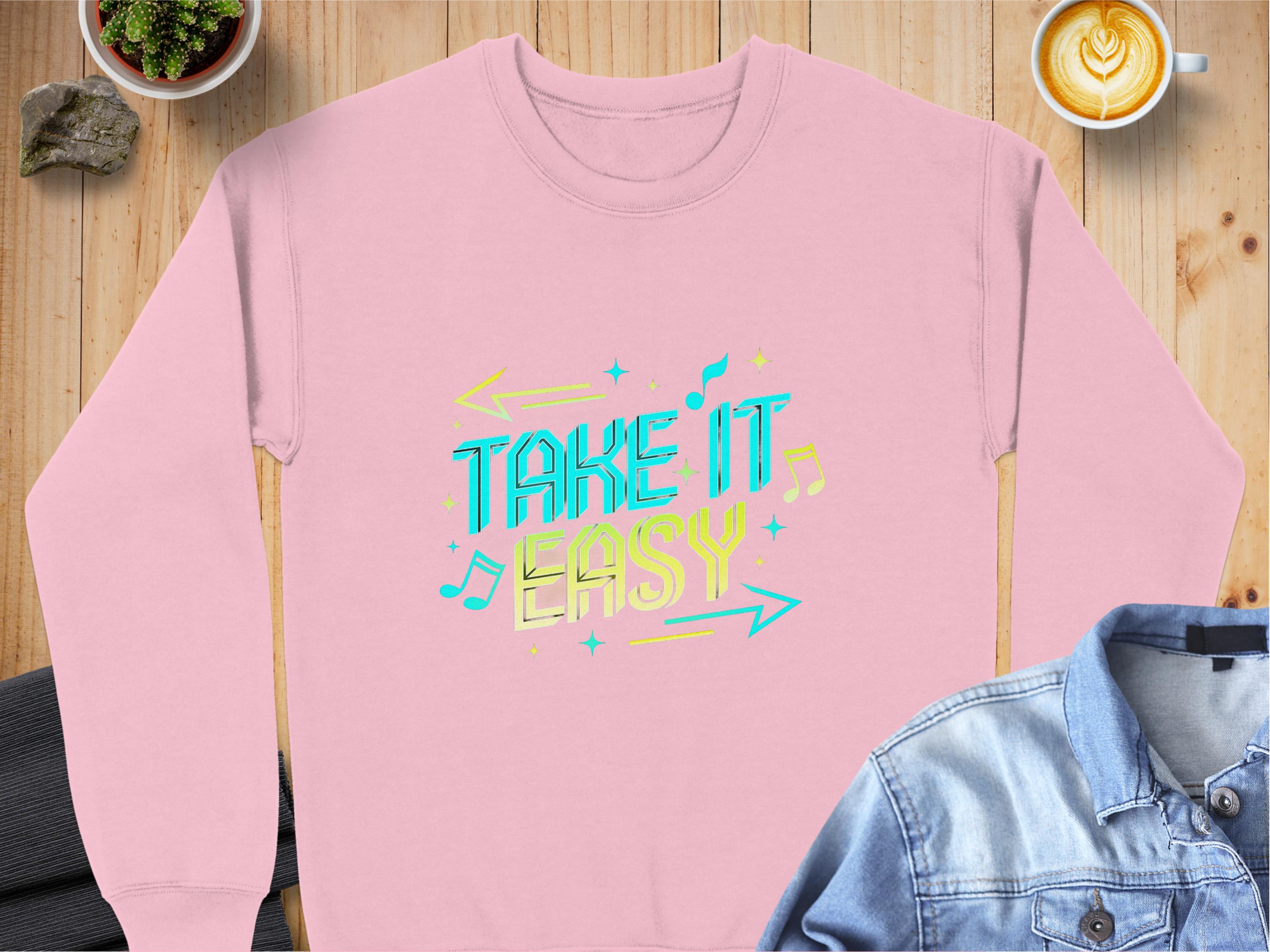 A pink Garment Graphics sweatshirt with Take It Easy in colorful retro-style text sits on a wooden table, surrounded by a latte, denim jacket, plant, and rock. The unisex fit and vibrant design make it ideal for anyone seeking style and comfort.