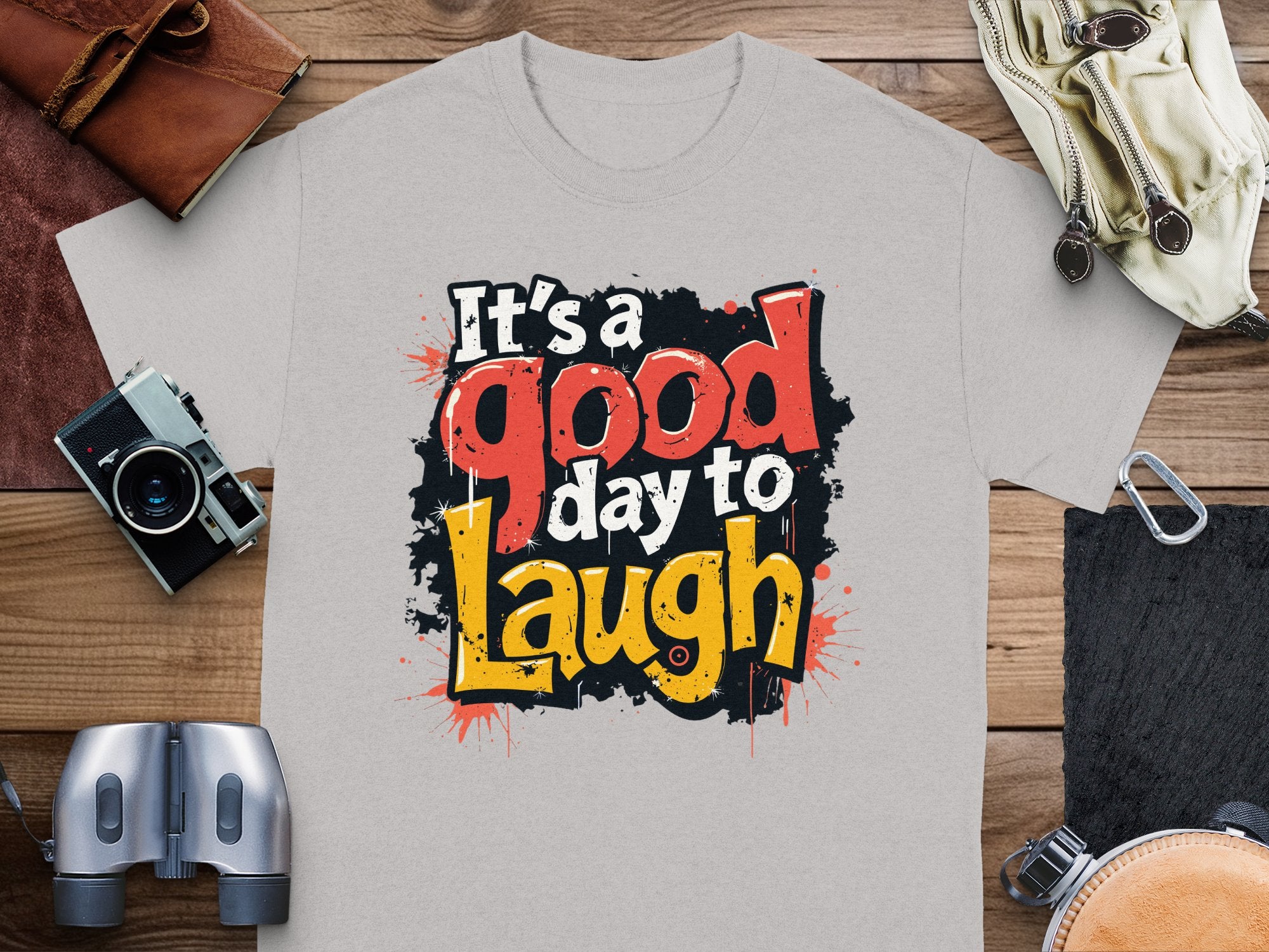 The Garment Graphics Its a Good Day to Laugh t-shirt is vibrant against its gray fabric. Positioned on wood, its paired with a camera, binoculars, leather pouch, and gloves—ideal for adventurers who live by its cheerful motto.